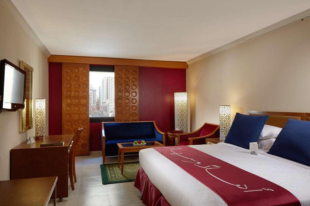 Makarem Ajyad Makkah Hotel Мекка Номер фото The photo showcases a hotel room featuring a modern design. In the center, there's a large bed with white linens and a decorative red runner. On either side of the bed, there are wooden nightstands with lamps. A blue sitting area is visible, composed