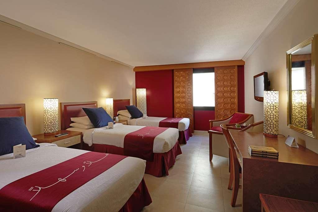 Makarem Ajyad Makkah Hotel Мекка Номер фото The photo shows a hotel room featuring three beds arranged with red and white bedding. The walls are painted in a neutral tone, and there are decorative lamps on nightstands beside each bed, providing ambient lighting. One of the walls has a window c