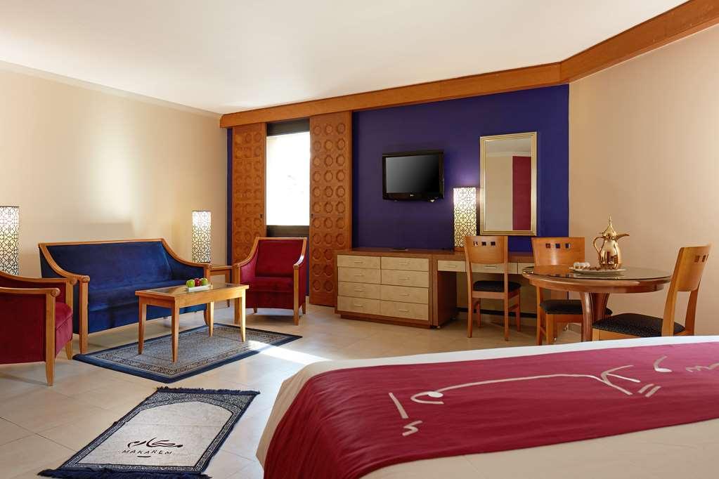 Makarem Ajyad Makkah Hotel Мекка Номер фото The image shows a hotel room with a modern, comfortable design. The room features a large bed with a red and white bedspread, a seating area that includes a blue sofa, two red chairs, and a wooden coffee table. There's a flat-screen TV mounted on the