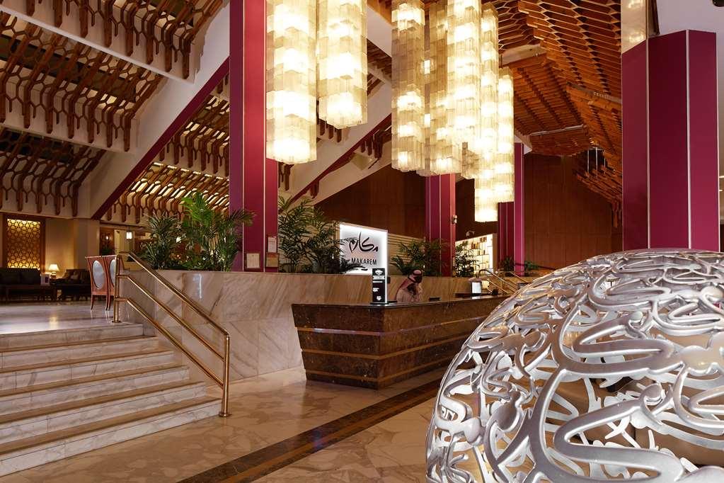 Makarem Ajyad Makkah Hotel Мекка Интерьер фото The photo depicts a luxurious hotel lobby. It features a grand entrance with a staircase leading up to a reception area. The lobby has high ceilings adorned with striking, modern light fixtures that hang down, providing a warm ambiance. The walls are