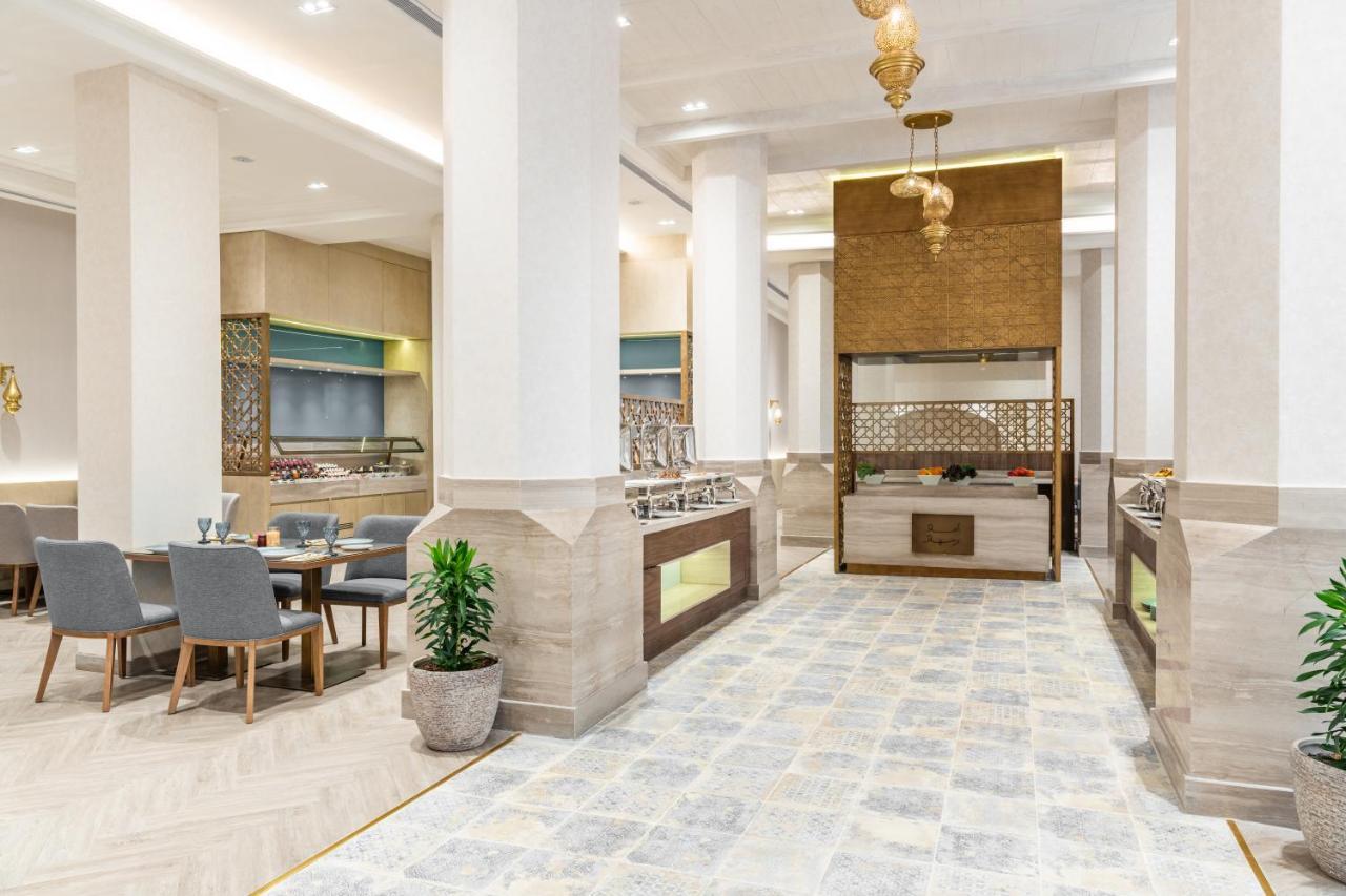 Makarem Ajyad Makkah Hotel Мекка Экстерьер фото The photo depicts a modern, elegant dining area, likely inside a restaurant or hotel. It features a spacious layout with tall ceilings and large columns. The flooring is a light-colored tiled design. 

To one side, there are several dining tables and