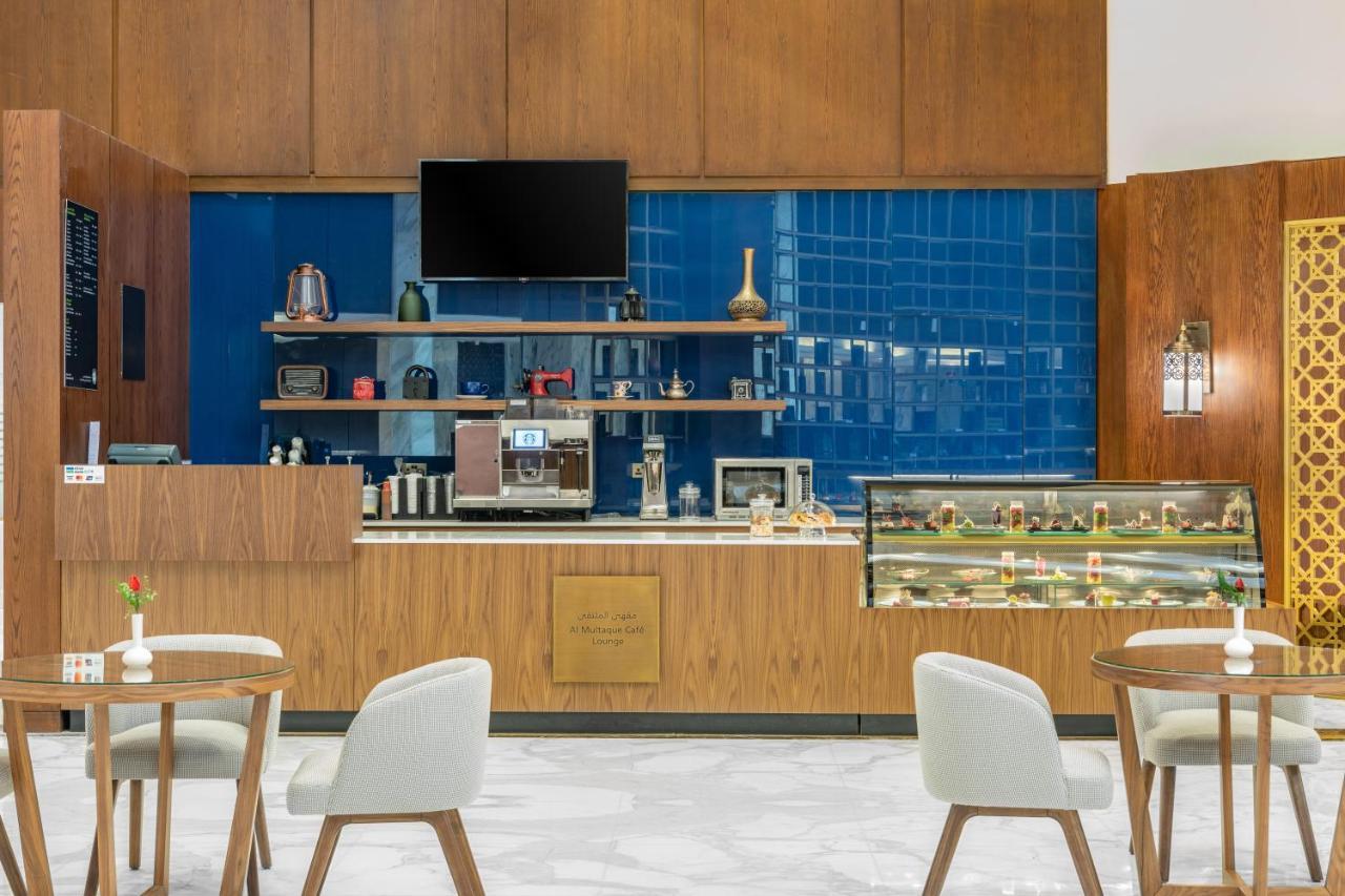 Makarem Ajyad Makkah Hotel Мекка Экстерьер фото The photo shows the interior of a café. The design features warm wood paneling and a striking blue tiled wall. In the foreground, there are several light-colored chairs and small tables arranged neatly. Behind the counter, there's a variety of coffee