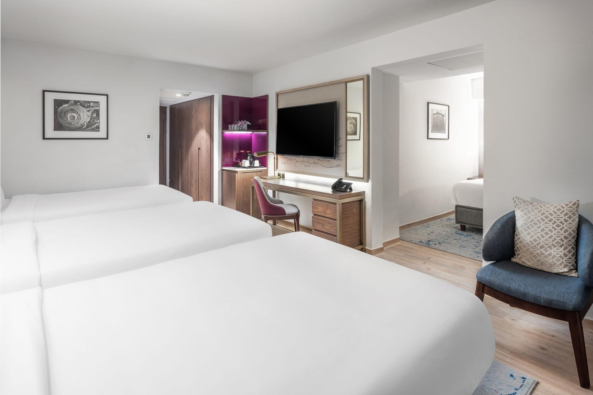 Makarem Ajyad Makkah Hotel Мекка Экстерьер фото The photo shows a modern hotel room featuring two white beds with smooth linens. The room has a minimalistic design with light-colored walls and wooden flooring. On one side, there is a desk and chair next to a television mounted on the wall, along w