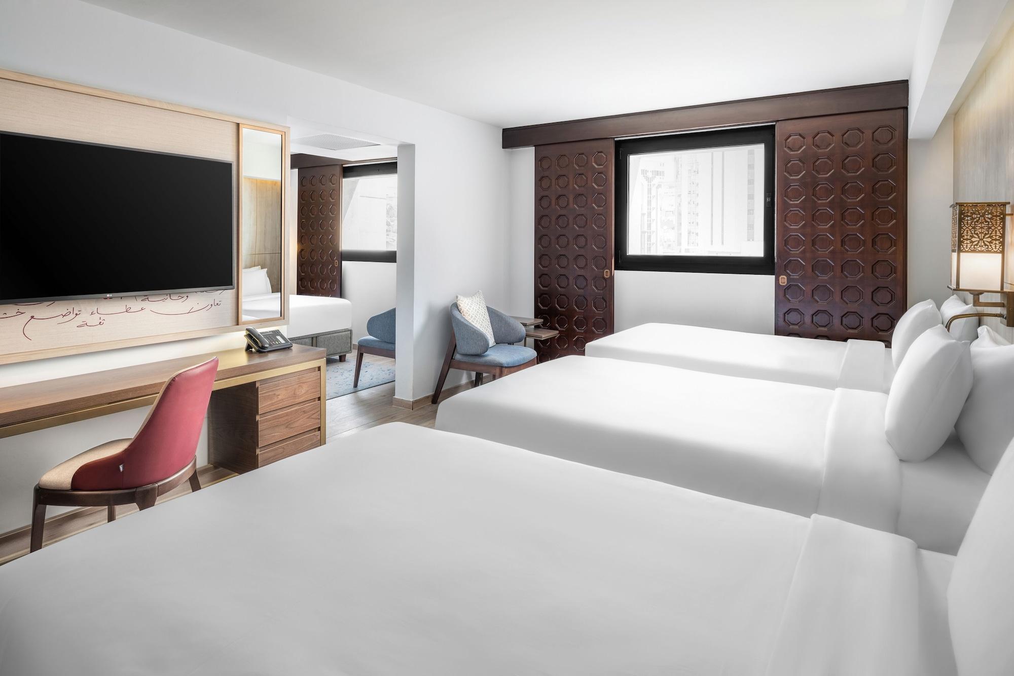 Makarem Ajyad Makkah Hotel Мекка Экстерьер фото The photo shows a hotel room featuring three beds with white linens. There is a large flat-screen TV mounted on the wall opposite the beds, and a wooden side table beneath it. A red chair is positioned near the TV, while a blue upholstered chair is p