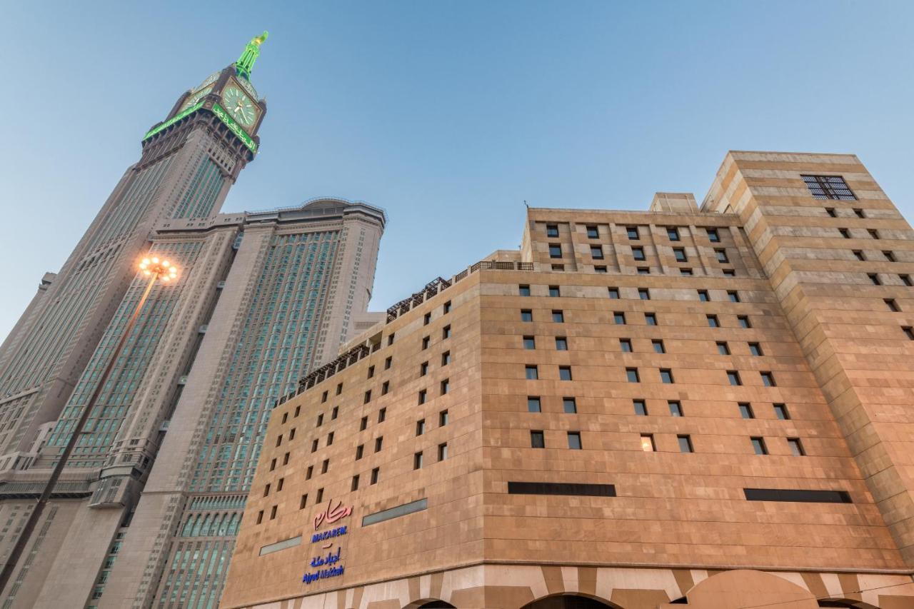 Makarem Ajyad Makkah Hotel Мекка Экстерьер фото The photo shows a prominent building with a modern architectural design, likely a hotel or commercial establishment, at the forefront. It features a light-colored stone facade with large windows. Behind it, there is a tall skyscraper, which has a dis